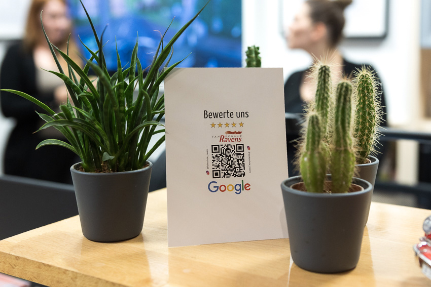 Zwei Kakteen mit einem Schild "bewerte uns bei Google" - Im Hintergrund zwei unscharfe Frauen im Gespräch.